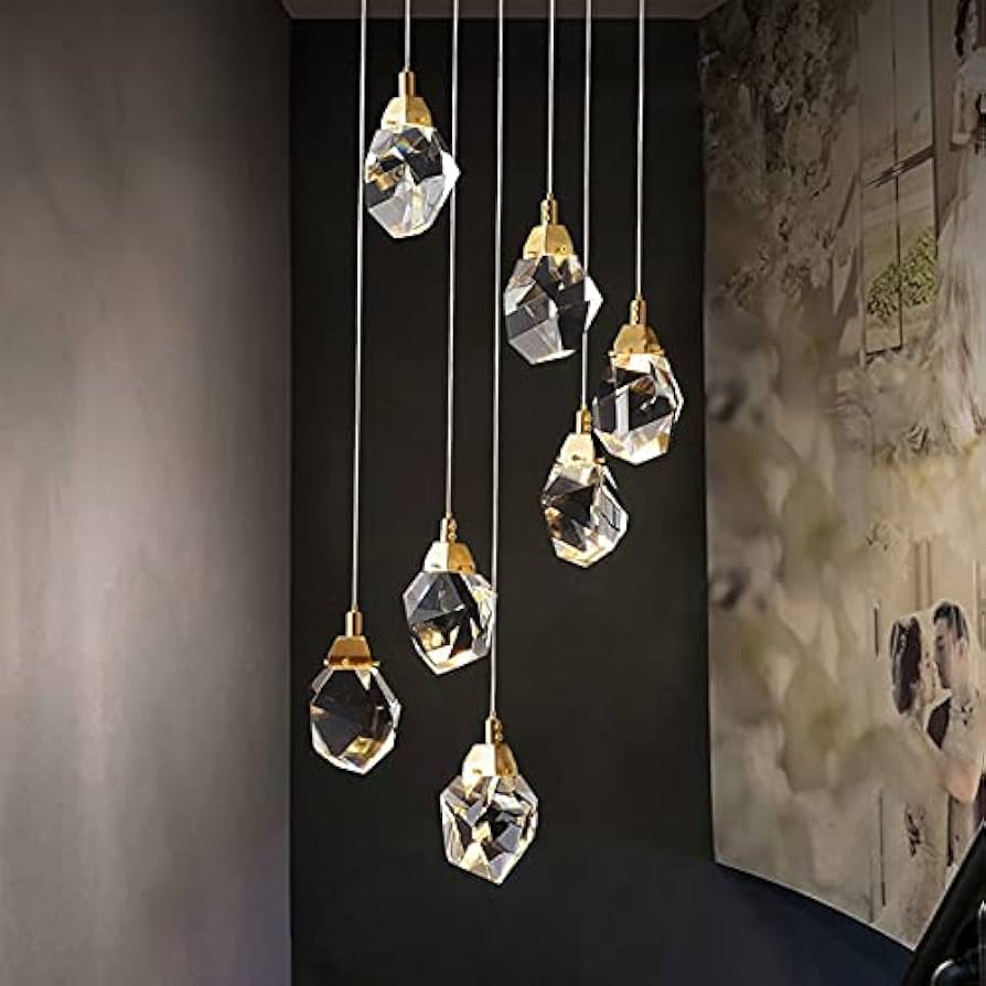 A set of modern geometric crystal pendant Astraea Cascade Duplex Chandelier with gold-toned caps and cords, hanging in a row against a dark wall next to a textured beige wall panel.