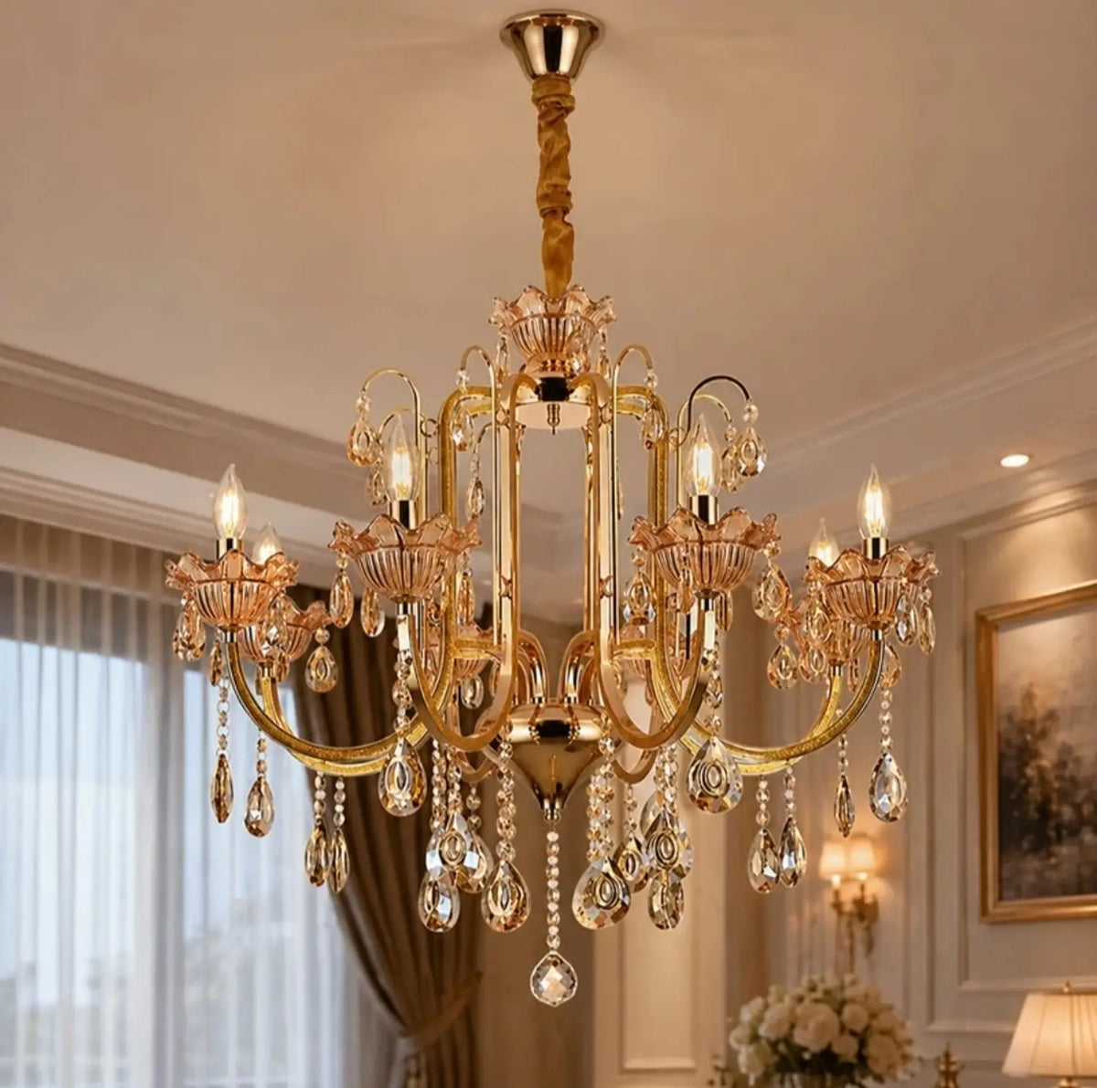 An ornate gold Luxe Wisteria Crystal Chandelier with multiple candle-style lights and hanging crystal droplets, shown installed in a decorated interior room with curtains, framed wall art, and warm ambient lighting.