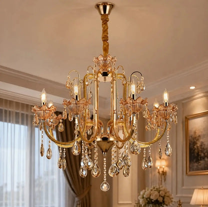 An ornate gold Luxe Wisteria Crystal Chandelier with multiple candle-style lights and hanging crystal droplets, shown installed in a decorated interior room with curtains, framed wall art, and warm ambient lighting.