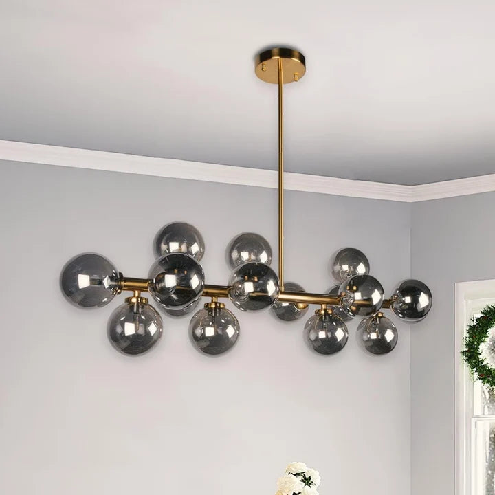 A modern Crystal Globe Dining Chandelier with a horizontal brass rod holding multiple spherical glass shades in a smoked gray finish, suspended from a ceiling in a room with light gray walls.