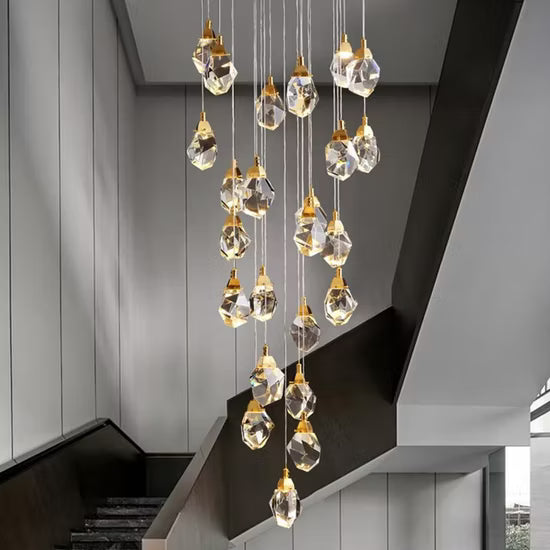 A modern multi-light crystal Astraea Cascade Duplex Chandelier with faceted clear glass teardrop pendants and gold-toned caps, hanging in a staggered waterfall arrangement over a contemporary staircase.