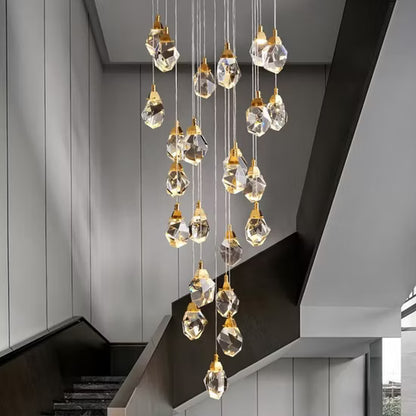 A modern multi-light crystal Astraea Cascade Duplex Chandelier with faceted clear glass teardrop pendants and gold-toned caps, hanging in a staggered waterfall arrangement over a contemporary staircase.