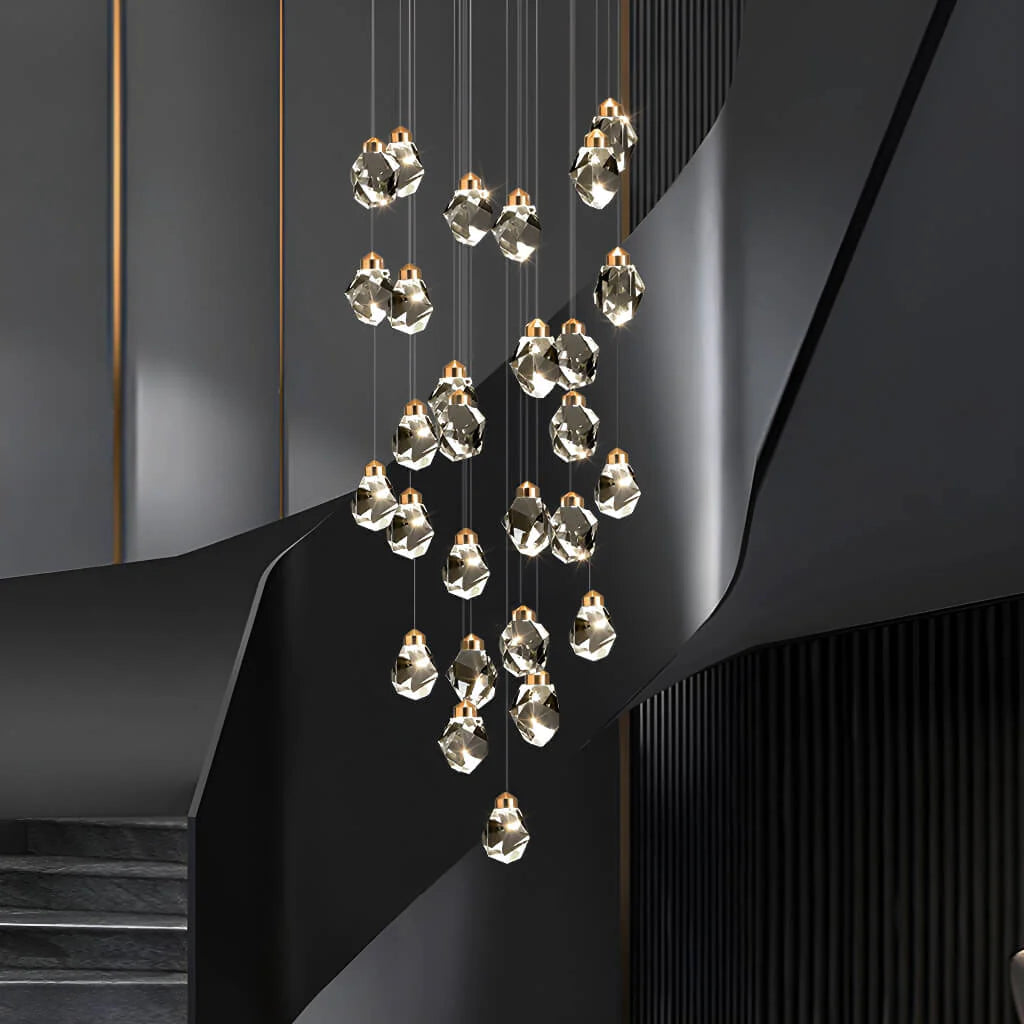A modern multi-light cascading Astraea Cascade Duplex Chandelier with faceted clear glass pendant bulbs and warm gold-toned caps, suspended in front of a sculptural dark gray staircase.