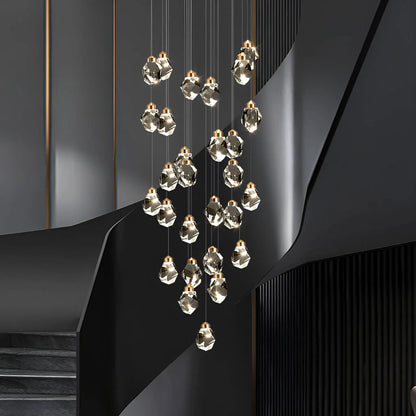 A modern multi-light cascading Astraea Cascade Duplex Chandelier with faceted clear glass pendant bulbs and warm gold-toned caps, suspended in front of a sculptural dark gray staircase.